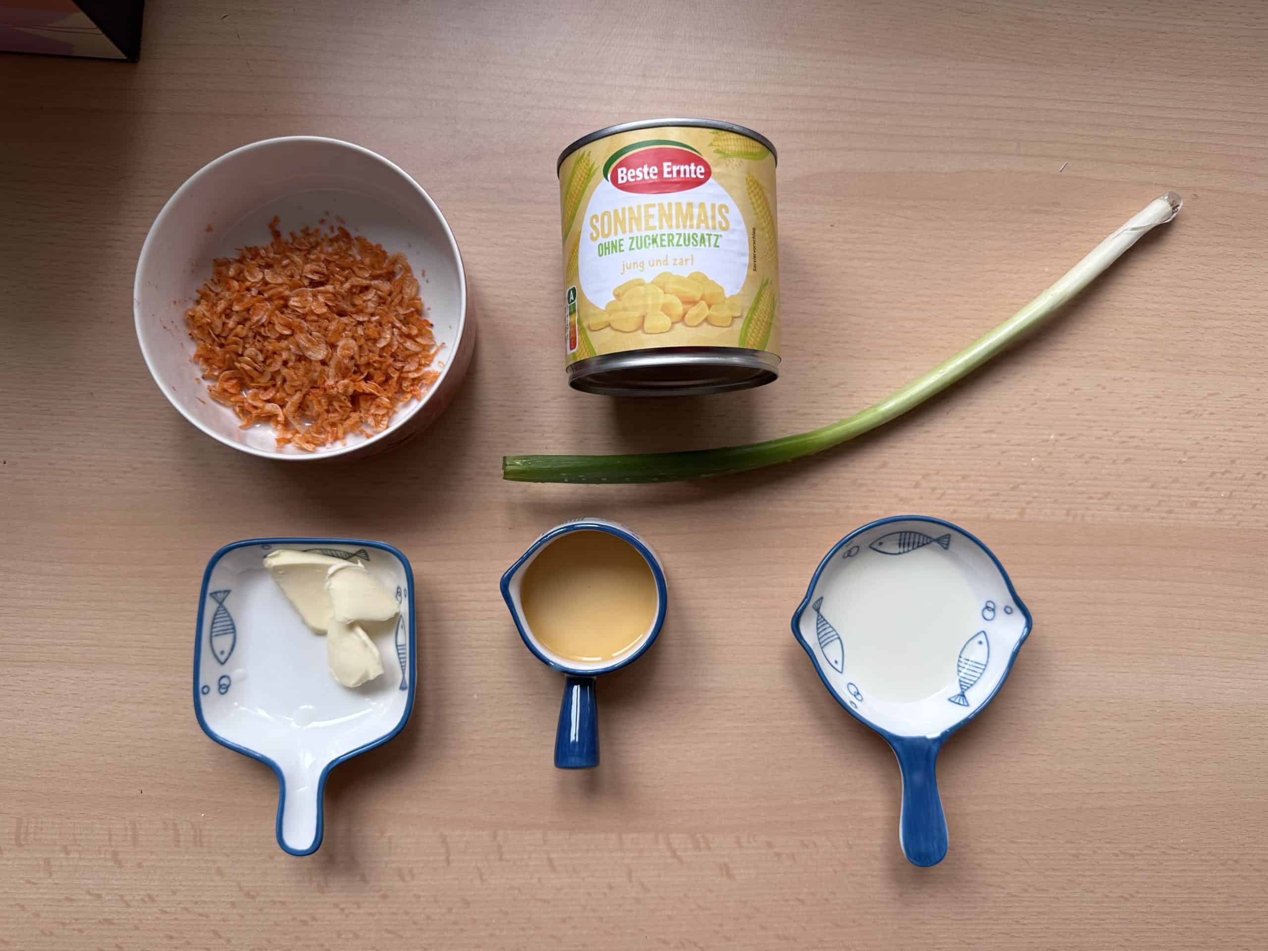 Bắp Xào - Vietnamesisch gebratener Mais mit Garnelen A photo showcasing the essential ingredients for Bap xao, a delicious Vietnamese stir-fried corn dish. The image features a can of corn kernels, fresh scallions, fish sauce, butter, and dried shrimps.