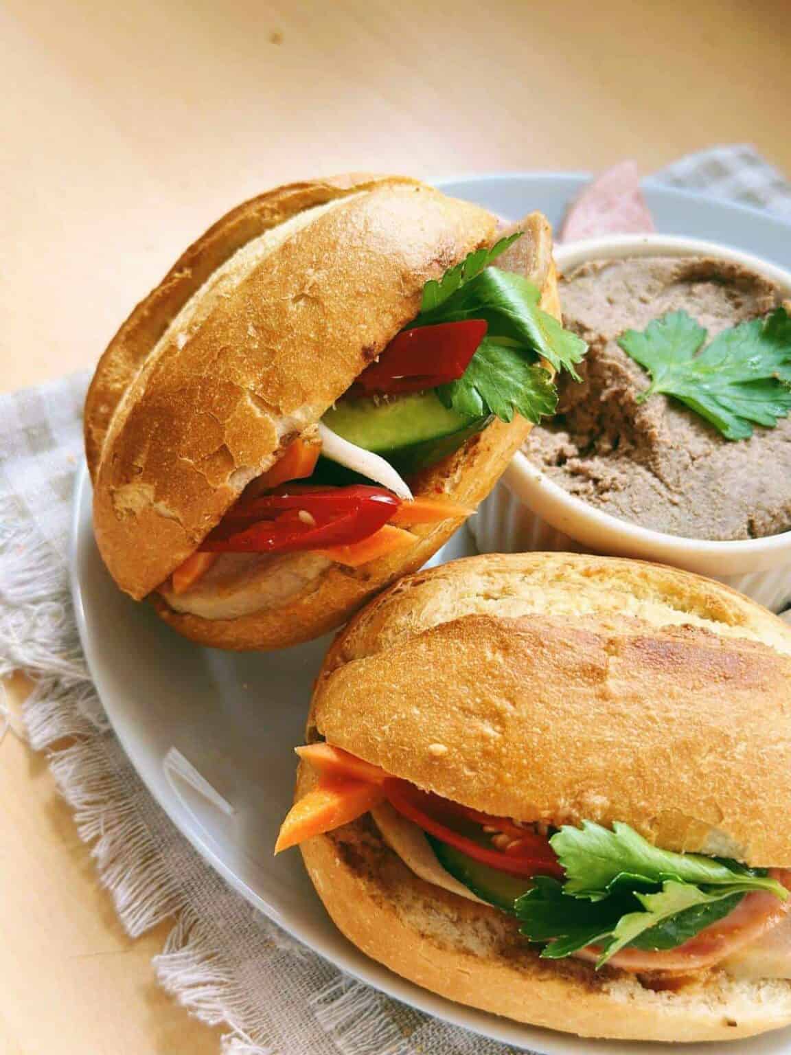 Close-up photo of a delicious Banh Mi Pate sandwich. The sandwich features a crusty baguette filled with layers of savory pate, fresh vegetables, cilantro, and pickled carrots.