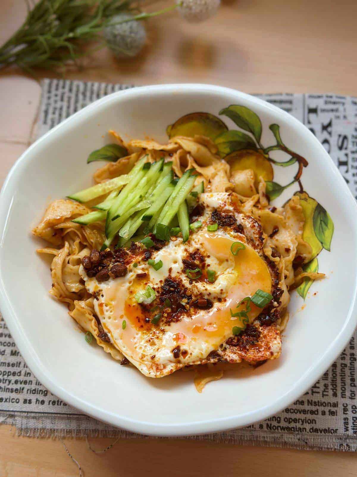 A close-up photo of a plate filled with steaming noodles topped with perfectly fried eggs in chili oil. The vibrant red chili oil glistens on the eggs, adding a spicy kick to the dish.