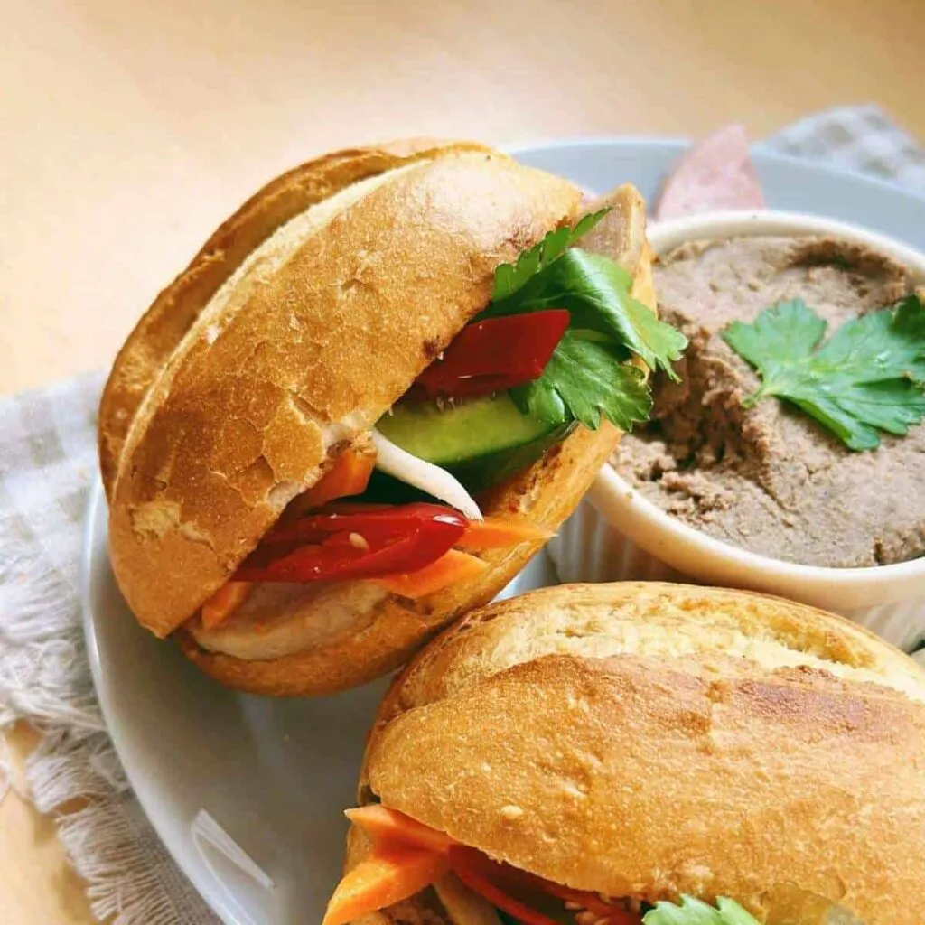 Vietnamese Spring Rolls with Peanut Sauce | Gỏi Cuốn Close-up photo of a delicious Banh Mi Pate sandwich. The sandwich features a crusty baguette filled with layers of savory pate, fresh vegetables, cilantro, and pickled carrots.