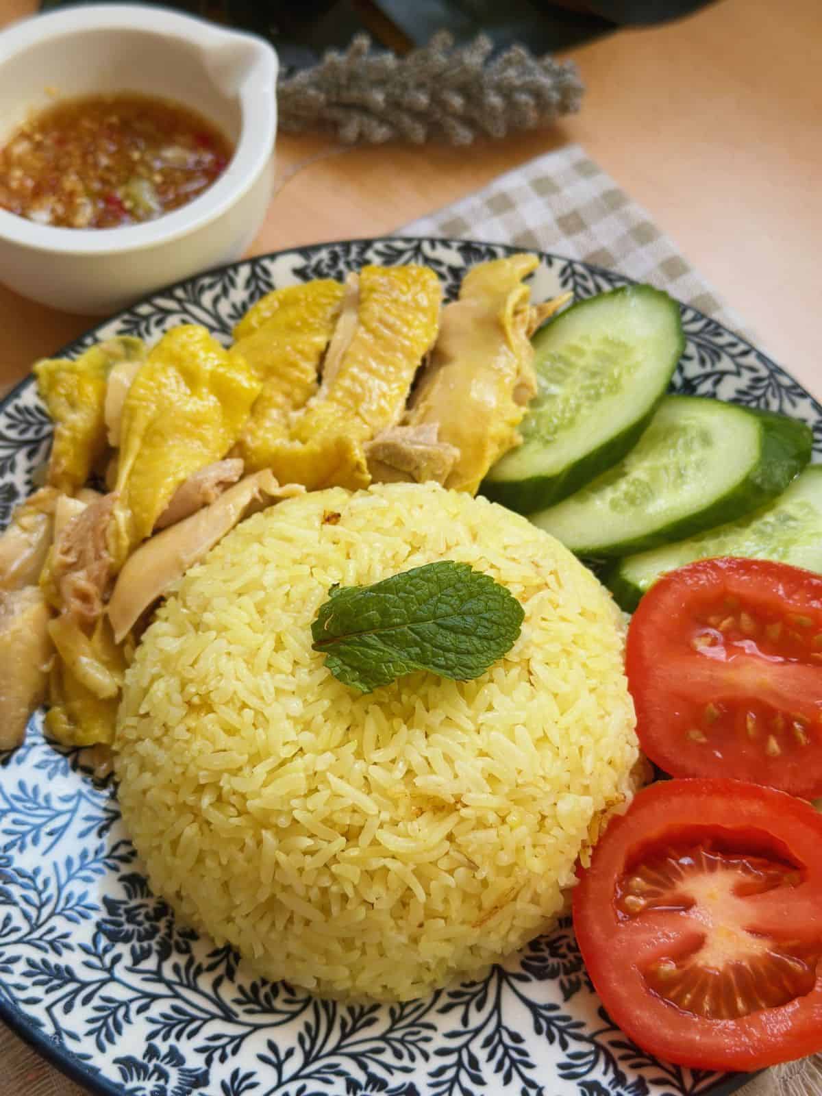 A delicious plate of Vietnamese Hainanese chicken rice with fragrant rice, tender chicken, and ginger fish sauce, garnished with herbs and served with cucumber and tomato slices