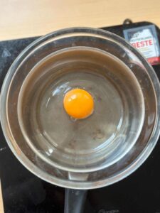 A glass bowl containing an egg yolk is positioned on top of a saucepan filled with boiling water, with the water level not touching the bowl.