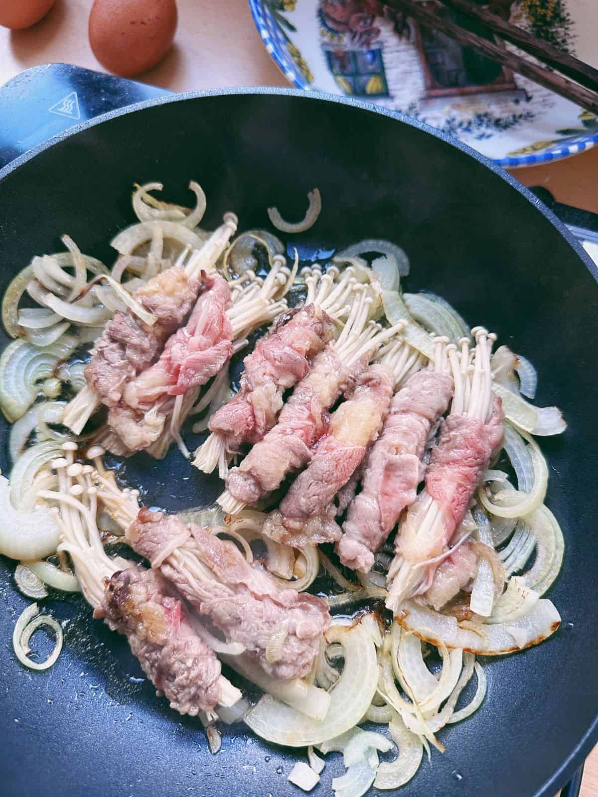Schnelle Rindfleischröllchen mit Enoki-Pilzen enoki beef rolls and sliced onion cooked in a pan.