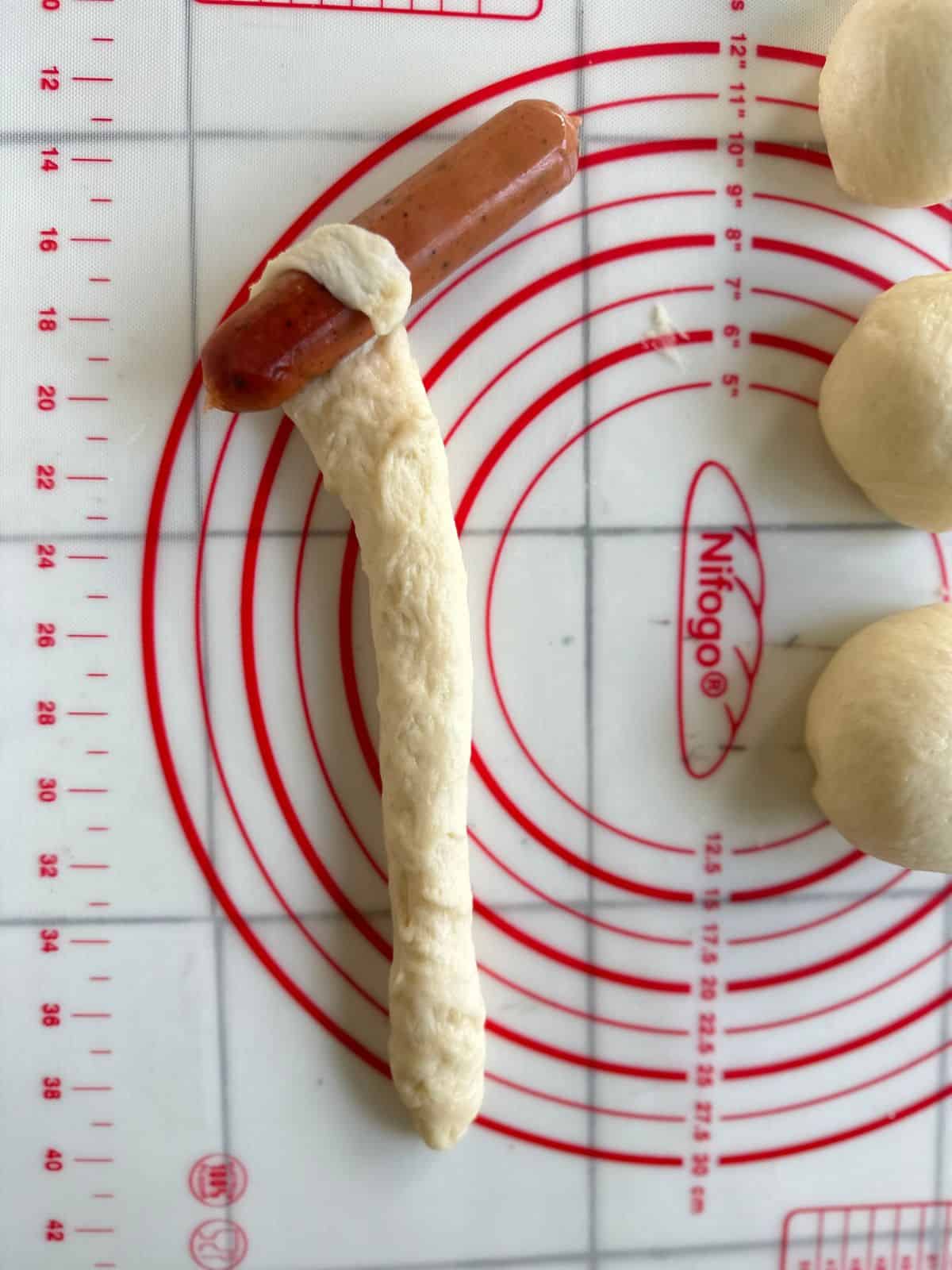 A long rope of dough being wrapped around one end of a hot dog, resting on a silicone pastry mat with measurement markings. Dough balls are visible nearby, ready to be shaped.