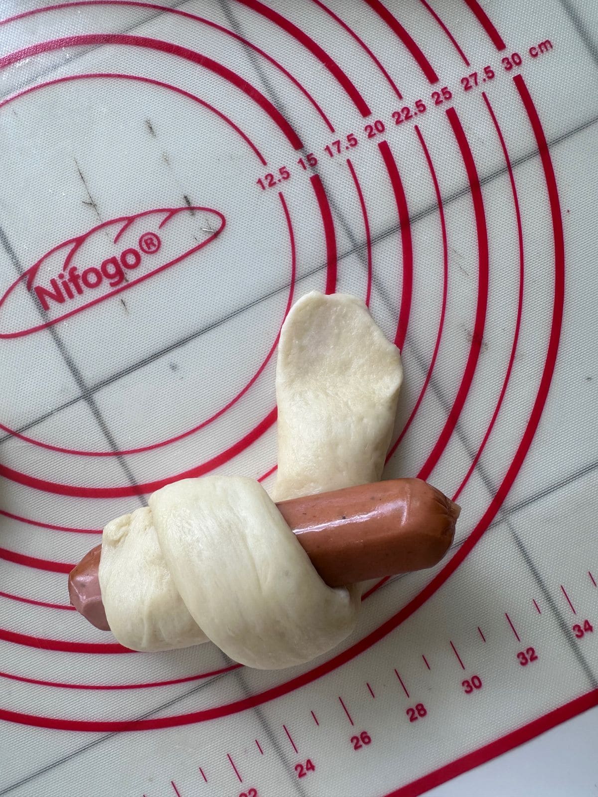 A hot dog partially wrapped in a spiral of soft dough, showing the shaping process of Chinese-style hot dog buns on a silicone baking mat.
