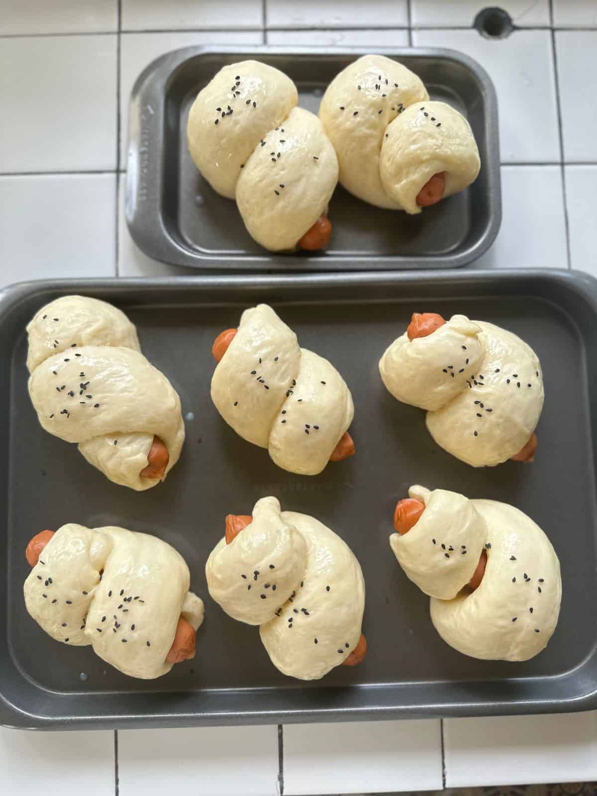 Unbaked Chinese hot dog buns arranged on two baking trays, brushed with egg wash and sprinkled with black sesame seeds, ready to go into the oven.
