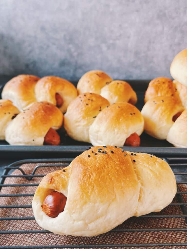 Freshly baked Chinese hot dog buns cooling on a wire rack. The buns are golden brown, soft, and topped with black sesame seeds, with the sausage peeking out at the ends.