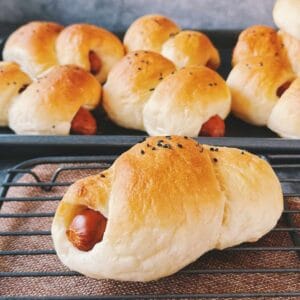 Freshly baked Chinese hot dog buns cooling on a wire rack. The buns are golden brown, soft, and topped with black sesame seeds, with the sausage peeking out at the ends.