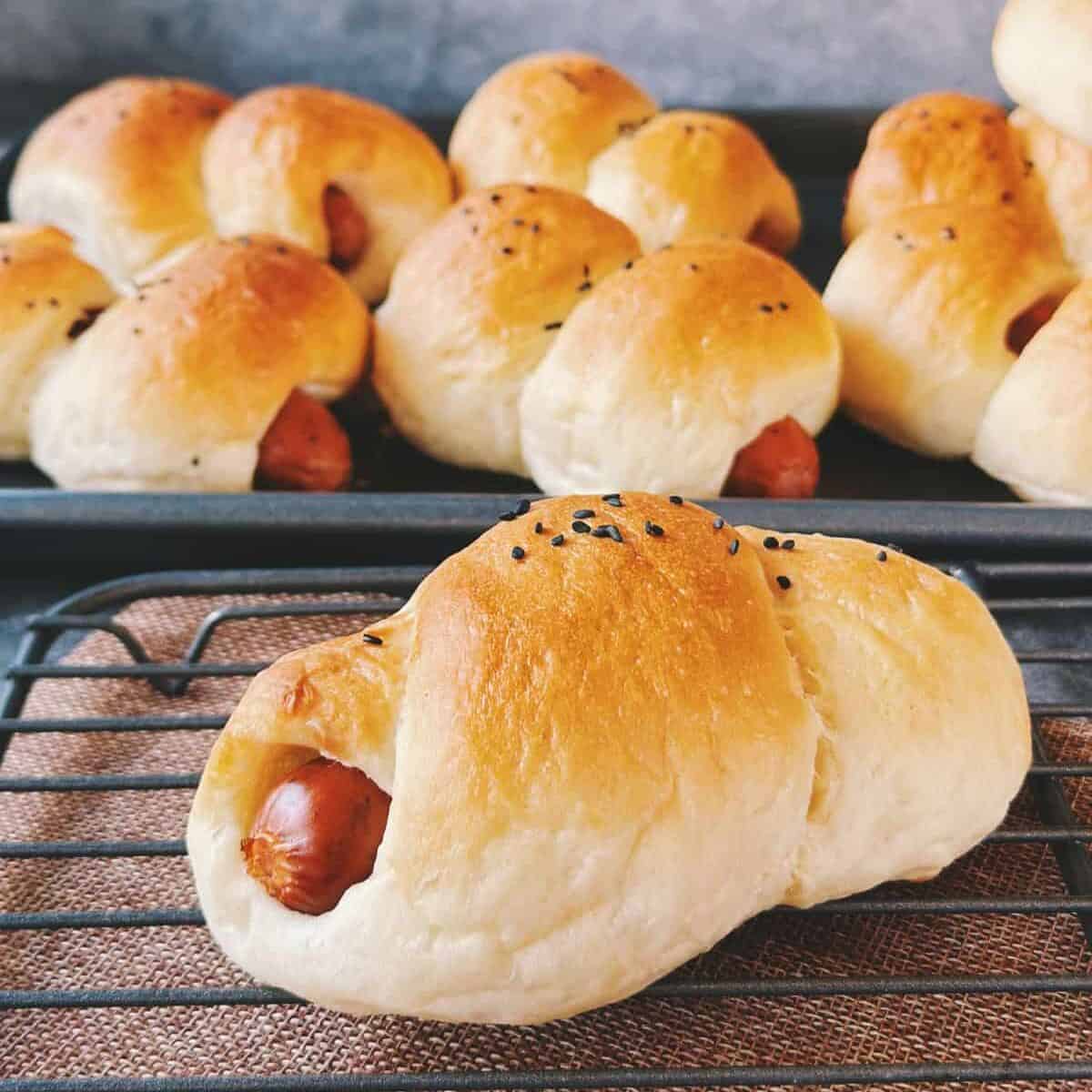 Freshly baked Chinese hot dog buns cooling on a wire rack. The buns are golden brown, soft, and topped with black sesame seeds, with the sausage peeking out at the ends.