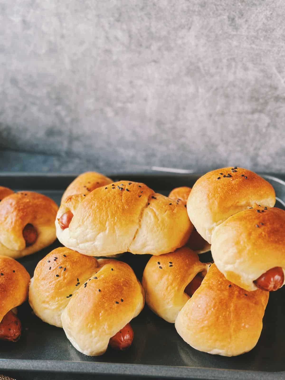 Freshly baked Chinese hot dog buns cooling on a wire rack. The buns are golden brown, soft, and topped with black sesame seeds, with the sausage peeking out at the ends.