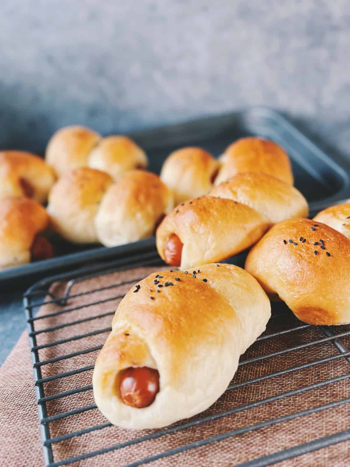 Freshly baked Chinese hot dog buns cooling on a wire rack. The buns are golden brown, soft, and topped with black sesame seeds, with the sausage peeking out at the ends.