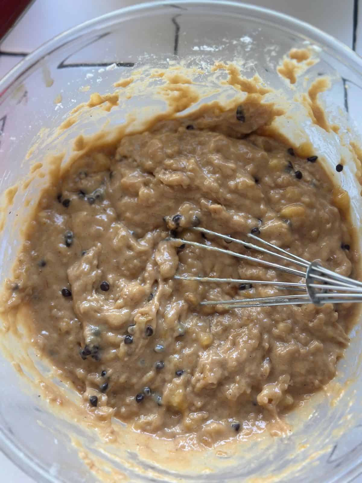 Eine Schüssel mit Bananenteig, der gerade mit einem Schneebesen vermischt wird. Kleine Luftblasen und ein paar dunkle Punkte von Schokostückchen sind sichtbar.