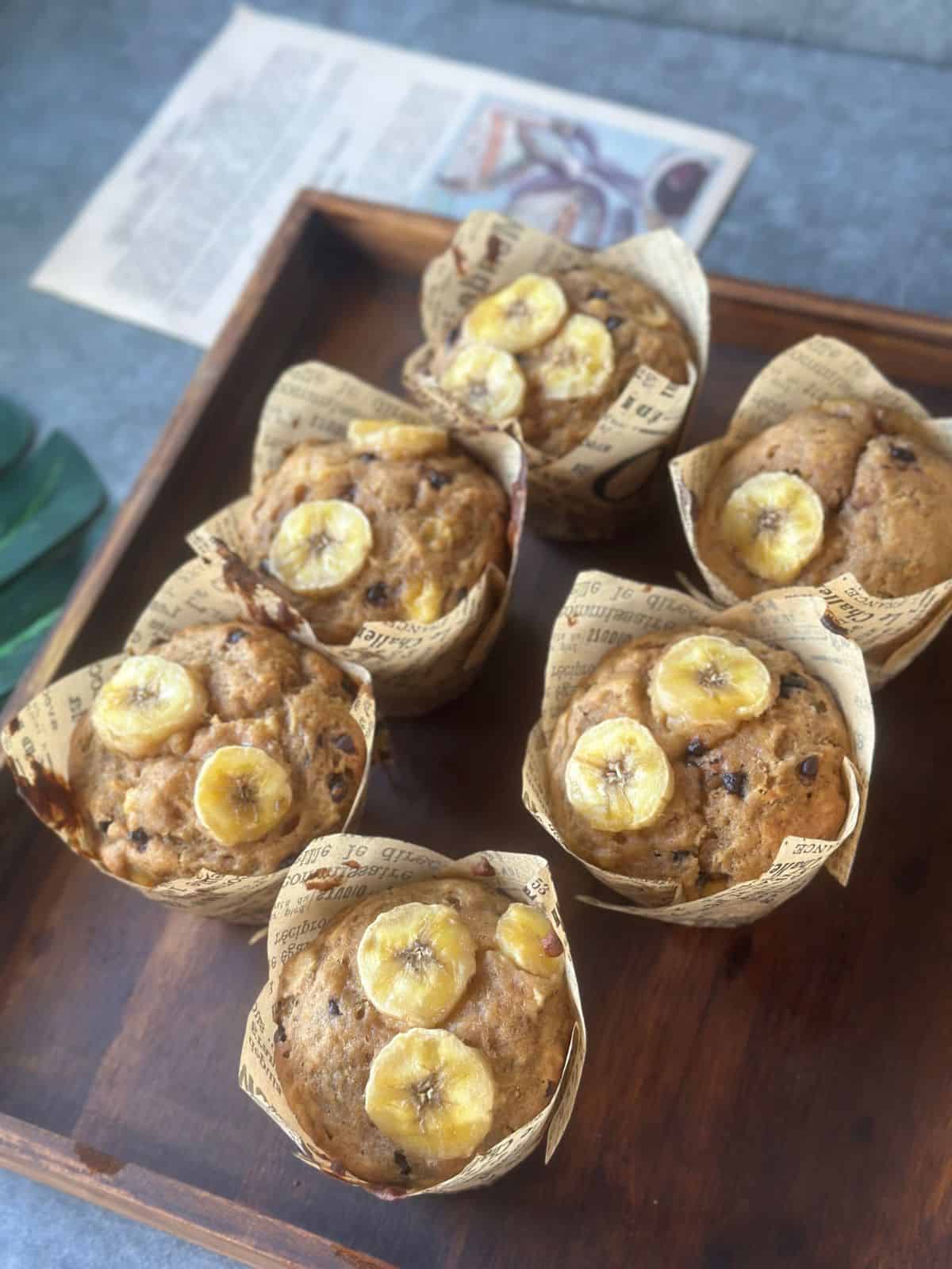 Fertig gebackene Bananenmuffins mit Schokostückchen, dekoriert mit Bananenscheiben, in braunem Papierförmchen auf einem Holztablett.