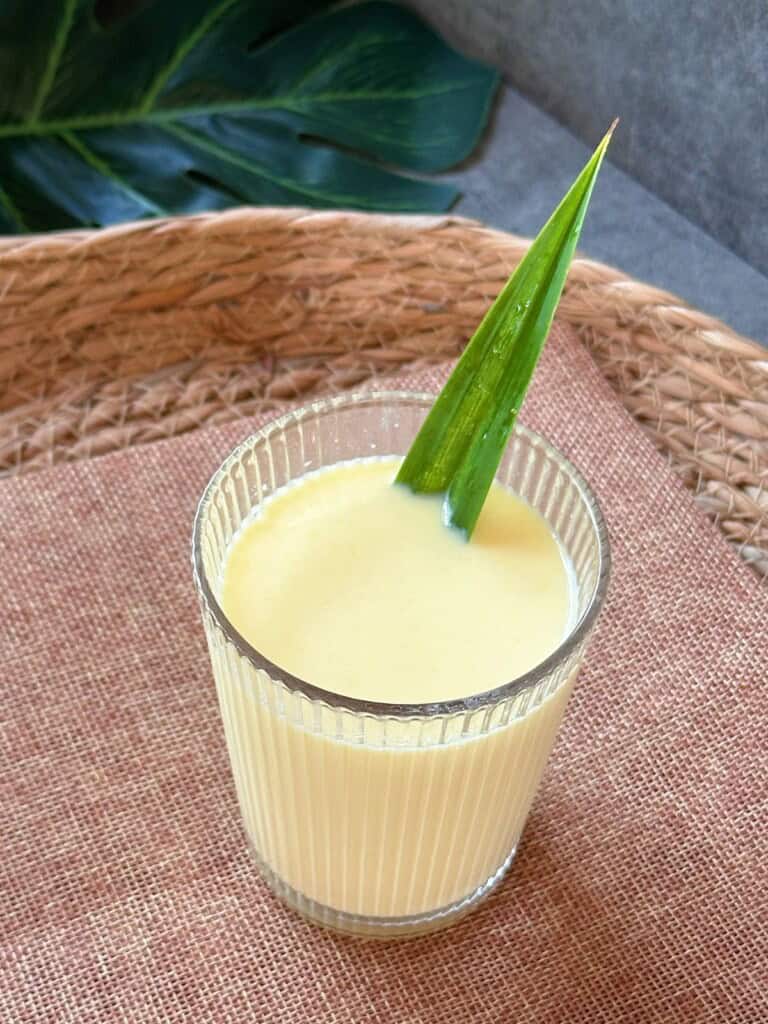 A glass of Vietnamese corn milk (Sữa Bắp) with a pandan leaf garnish, placed on a woven tray.