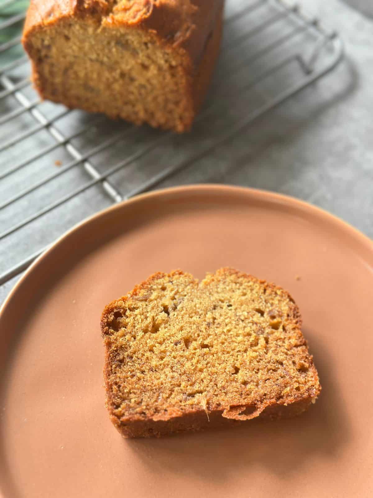 Bester Bananenkuchen – Saftig & Einfach Gelingsicher Eine Scheibe Bananenkuchen auf einem beigefarbenen Teller, im Hintergrund der restliche Kuchen auf einem Kuchengitter.