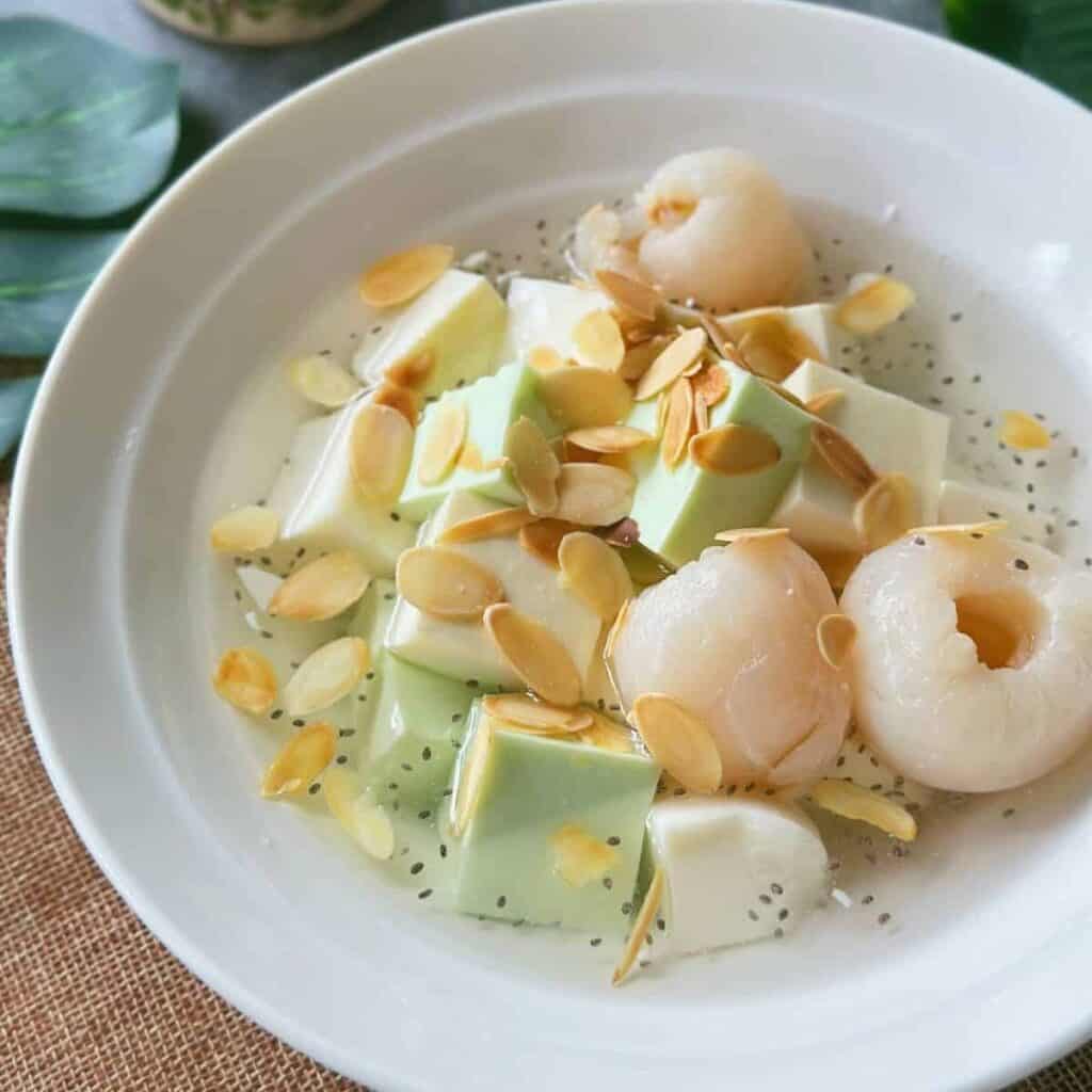 Bạc Xỉu (The Iconic Saigon's White Coffee) A bowl of Chè Khúc Bạch with white and green pudding cubes, lychees, basil seeds, and toasted almond slices in chilled syrup.