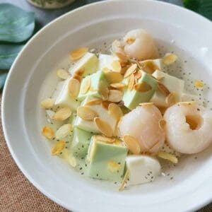 Chè Khúc Bạch – Vietnamese Panna Cotta Dessert (Easy!) A bowl of Chè Khúc Bạch with white and green pudding cubes, lychees, basil seeds, and toasted almond slices in chilled syrup.