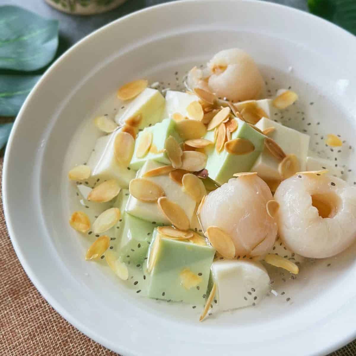 A bowl of Chè Khúc Bạch with white and green pudding cubes, lychees, basil seeds, and toasted almond slices in chilled syrup.
