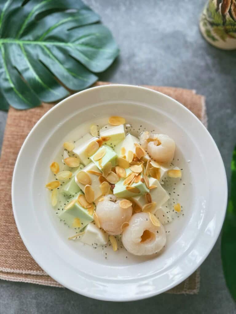A bowl of Chè Khúc Bạch with white and green pudding cubes, lychees, basil seeds, and toasted almond slices in chilled syrup.