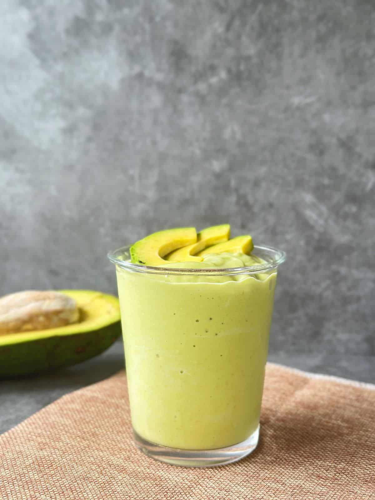 A creamy Vietnamese avocado smoothie in a clear glass, topped with fresh avocado slices, with half an avocado and seed in the background on a textured mat.