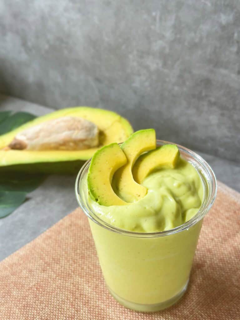 Close-up of a thick Vietnamese avocado smoothie in a clear glass, garnished with avocado slices, with a halved avocado in the background.