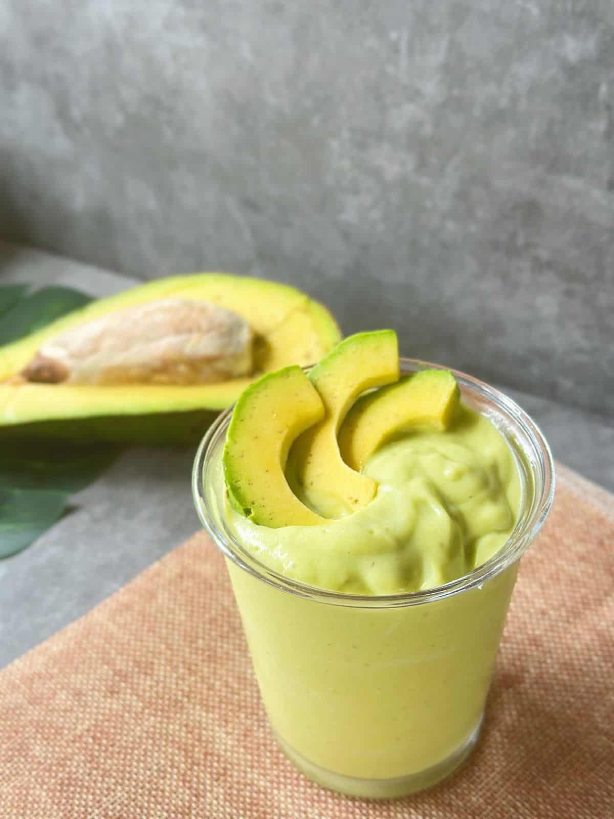 Close-up of a thick Vietnamese avocado smoothie in a clear glass, garnished with avocado slices, with a halved avocado in the background.