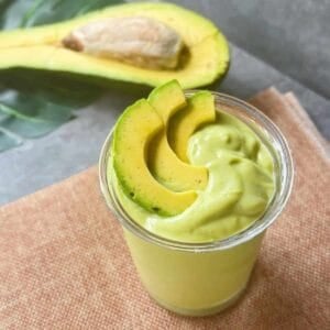 Top view of a rich Vietnamese avocado smoothie with avocado slices on top, placed on a textured mat, with a halved avocado in the background.