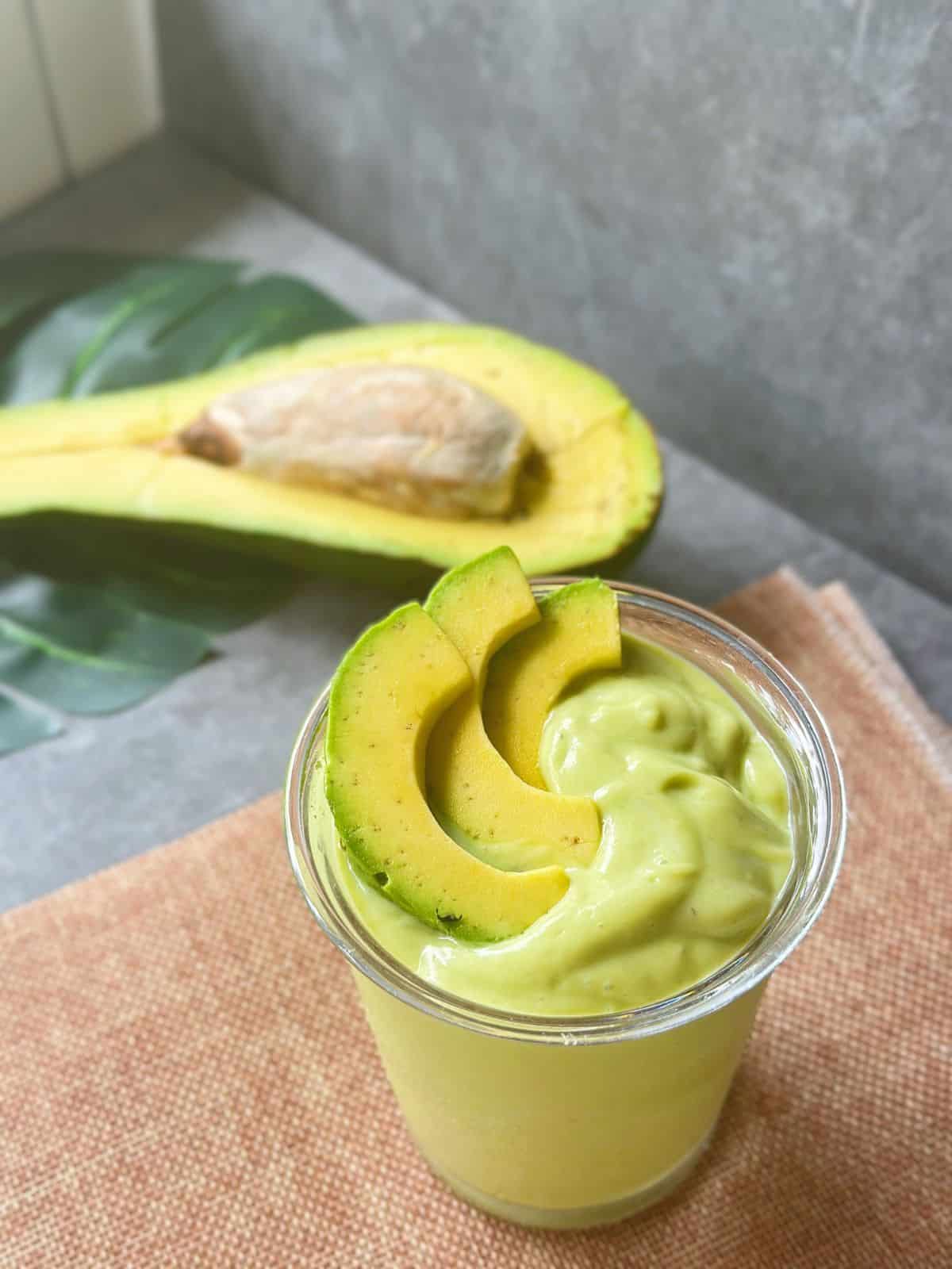 Top view of a rich Vietnamese avocado smoothie with avocado slices on top, placed on a textured mat, with a halved avocado in the background.