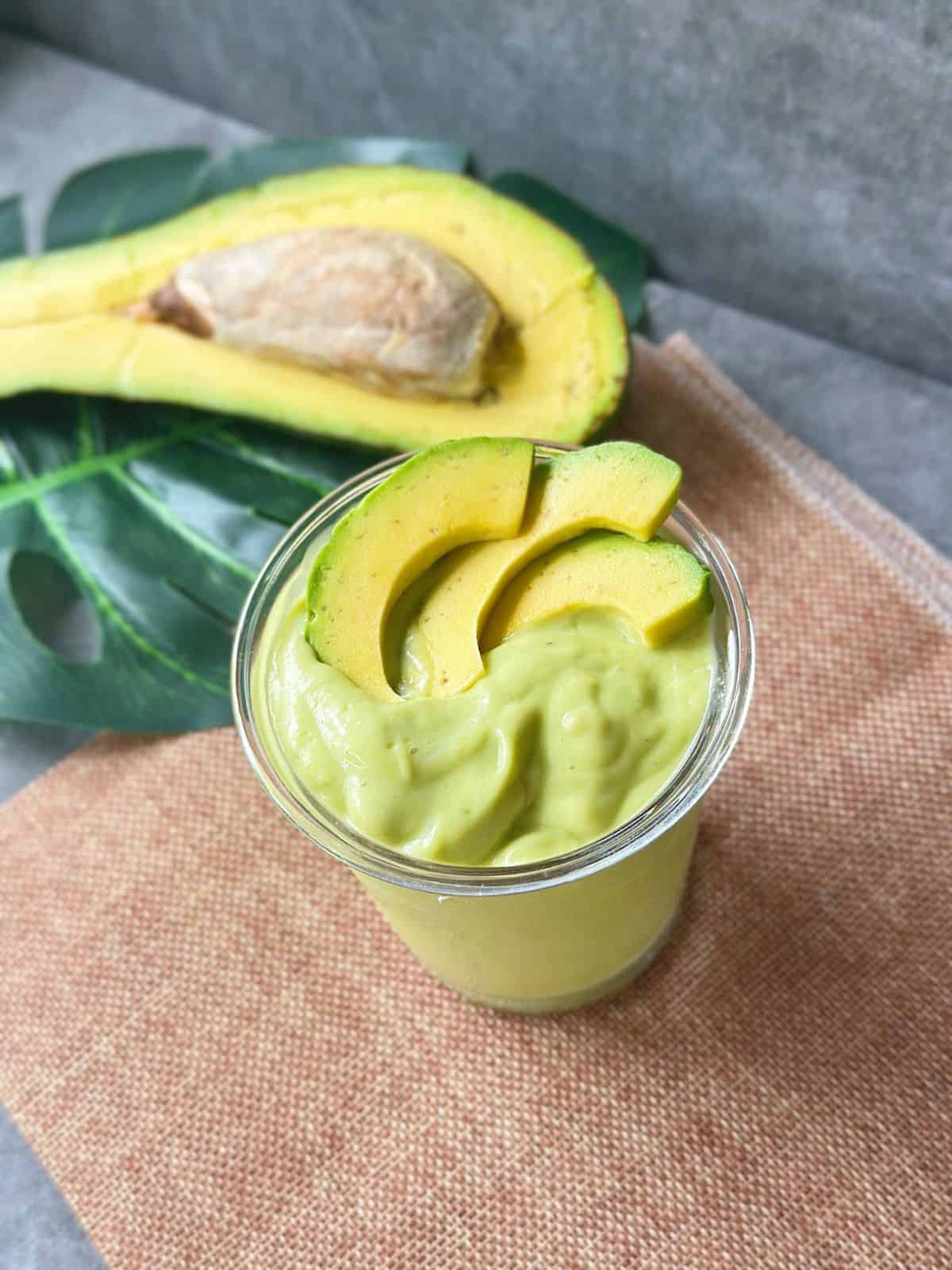 Overhead shot of a Vietnamese avocado smoothie in a clear glass, decorated with avocado slices, with half an avocado resting on a green leaf in the background.