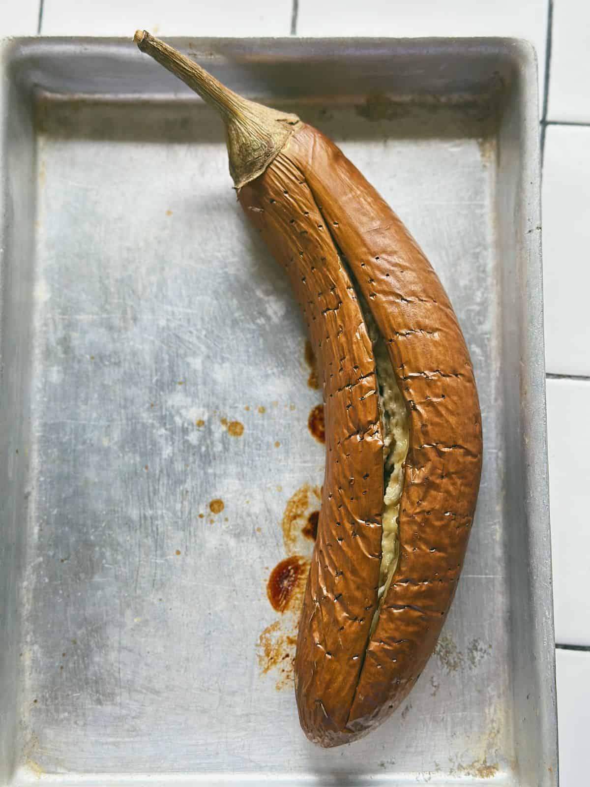 Whole eggplant baked until the skin is wrinkled and split, resting on a baking tray.