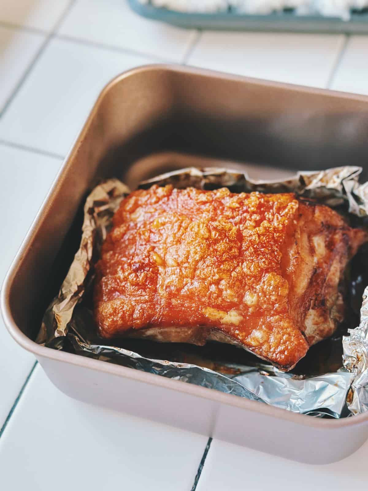 A piece of golden-brown crispy pork belly resting in a baking tray lined with crumpled aluminum foil, showcasing blistered, crunchy skin.