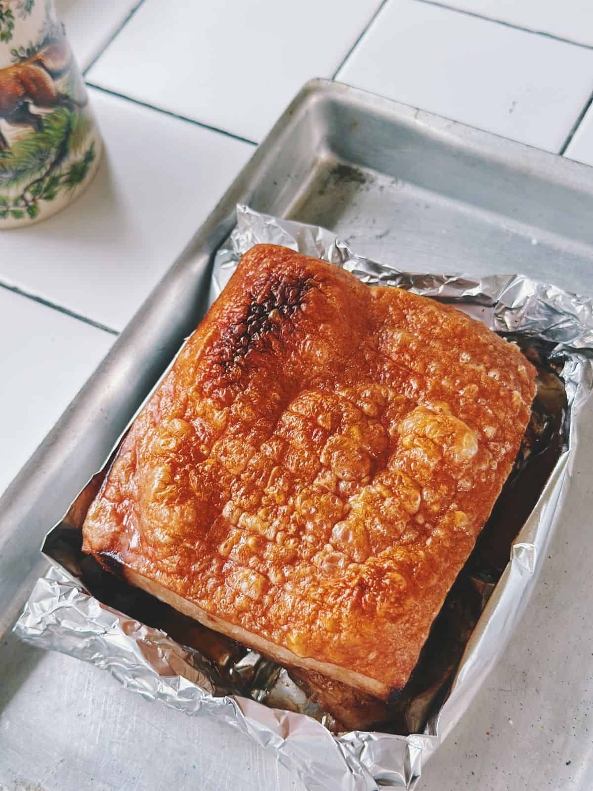 Close-up of roasted pork belly with puffed, crispy skin in a metal baking tray, with one small darkened spot from broiling.