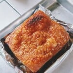 Angle shot of crackly-skinned pork belly sitting in foil-lined tray, with crispy bubbles across the surface indicating perfect roasting.