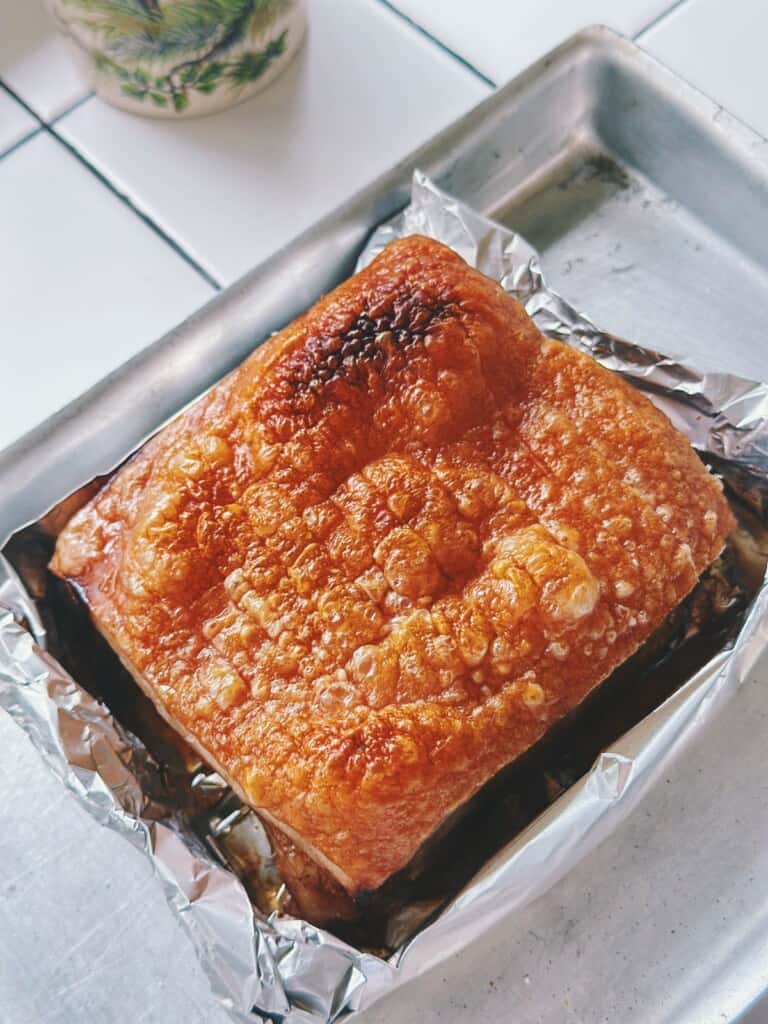 Angle shot of crackly-skinned pork belly sitting in foil-lined tray, with crispy bubbles across the surface indicating perfect roasting.