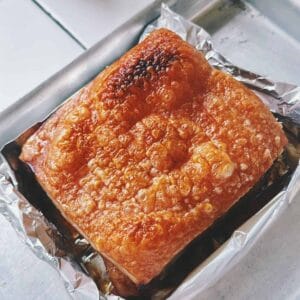 Angle shot of crackly-skinned pork belly sitting in foil-lined tray, with crispy bubbles across the surface indicating perfect roasting.