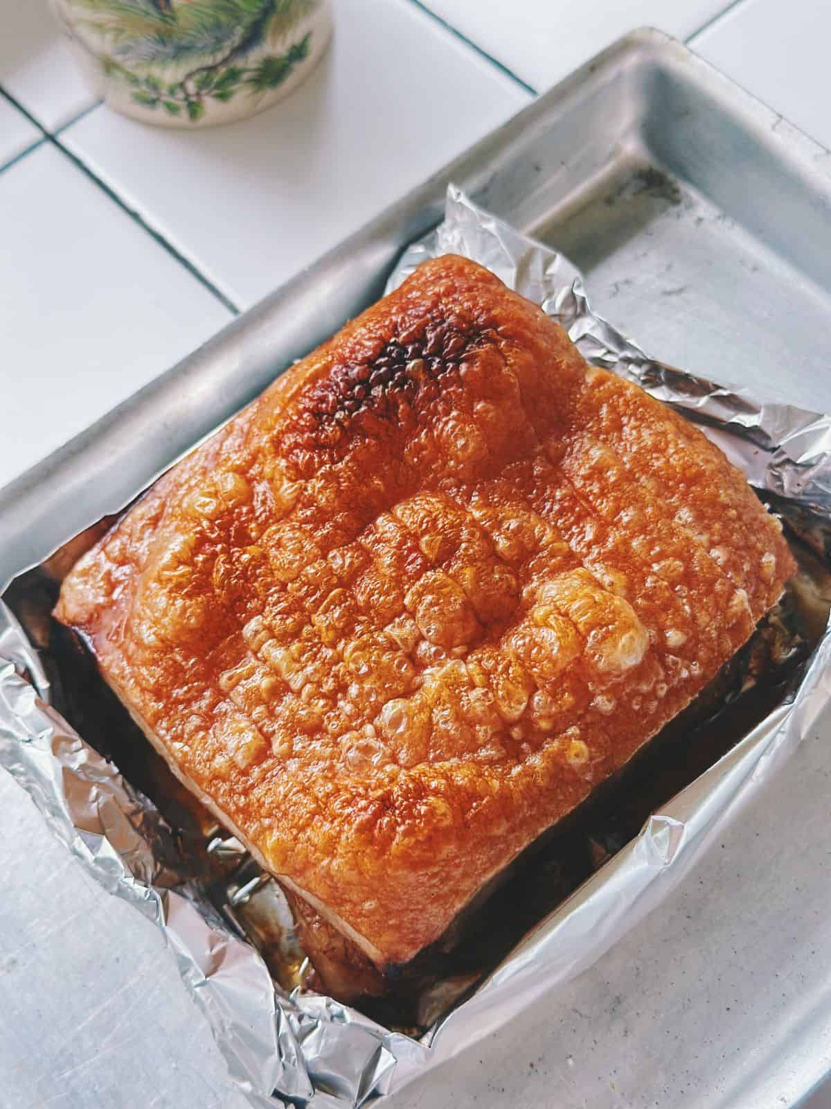 Angle shot of crackly-skinned pork belly sitting in foil-lined tray, with crispy bubbles across the surface indicating perfect roasting.