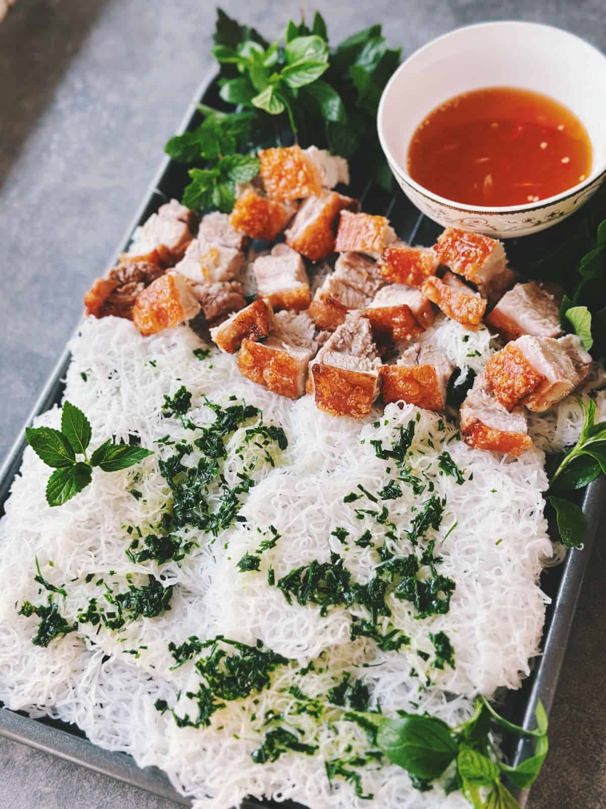 A tray of Vietnamese-style crispy pork belly slices served with steamed rice vermicelli sheets (bánh hỏi), fresh herbs, and a bowl of dipping sauce (nước chấm).