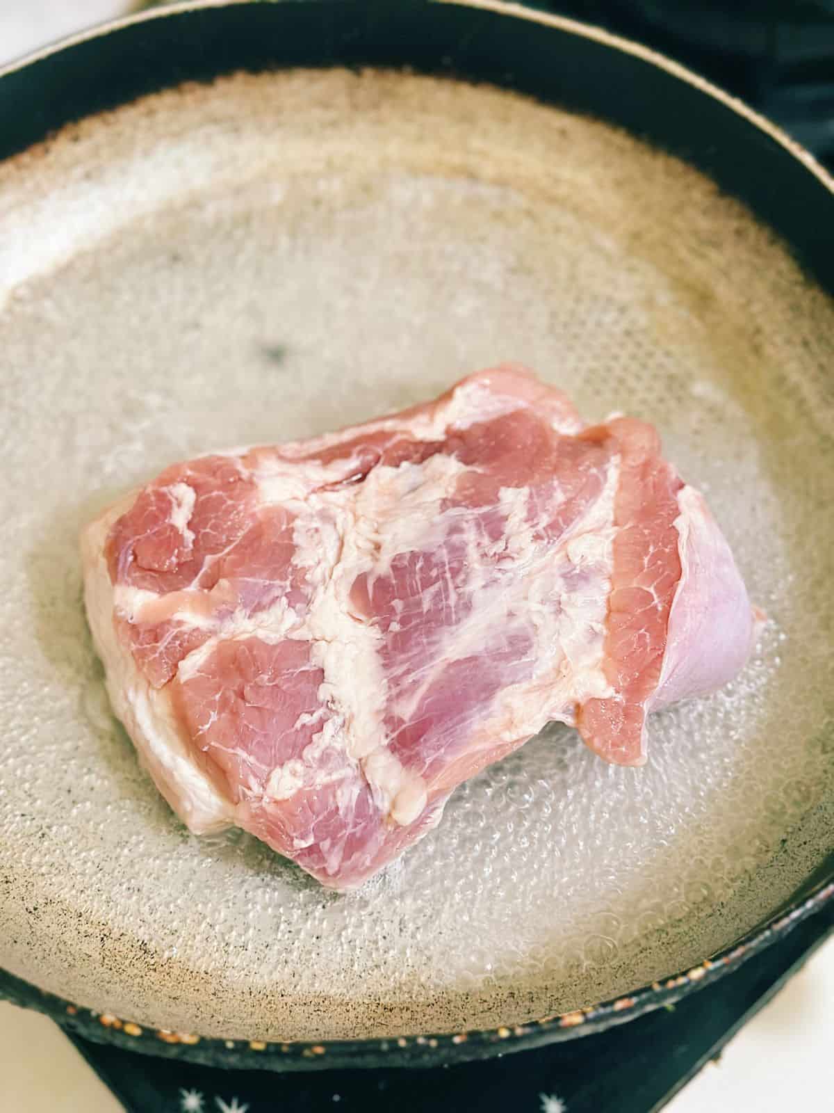A slab of raw pork belly being blanched skin-side up in a shallow pan of boiling water to help soften the skin and remove odors.