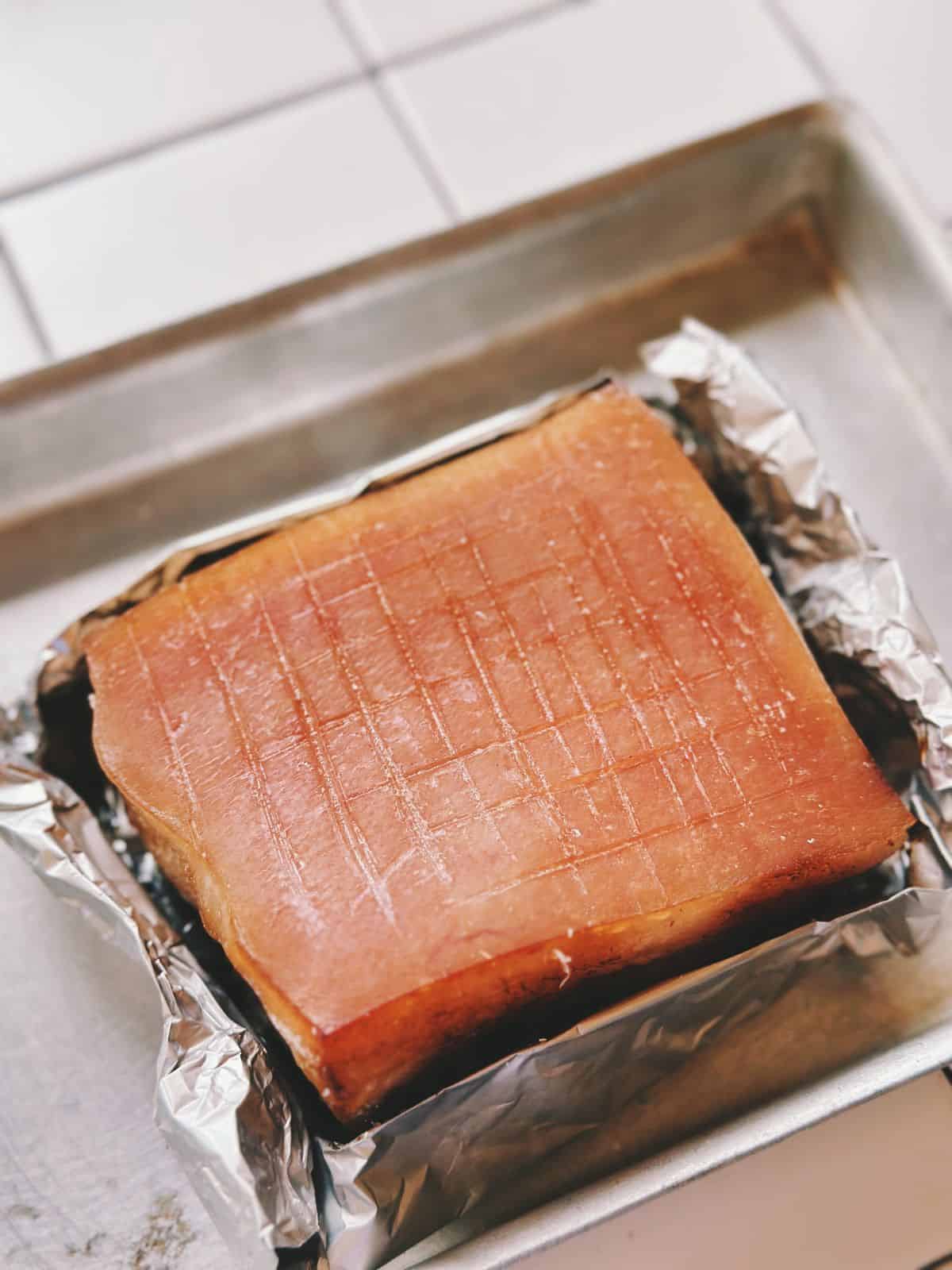 The pork belly placed skin-side up on a tray, scored in a criss-cross pattern and wrapped in foil around the meat side.