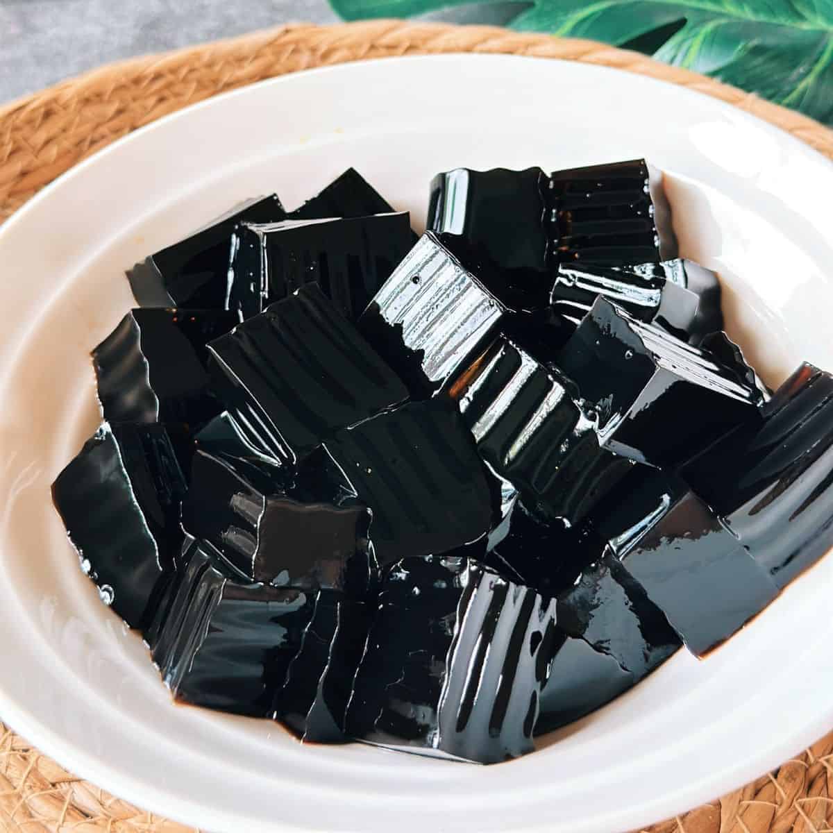 Close-up of glossy black grass jelly cubes served in a white bowl.