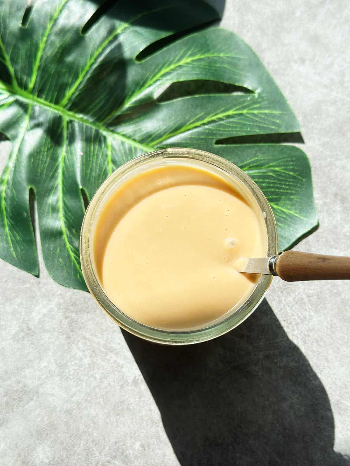 A jar of Hainanese Kaya coconut jam with a glossy, golden-brown texture, placed on a wooden coaster against a neutral background.