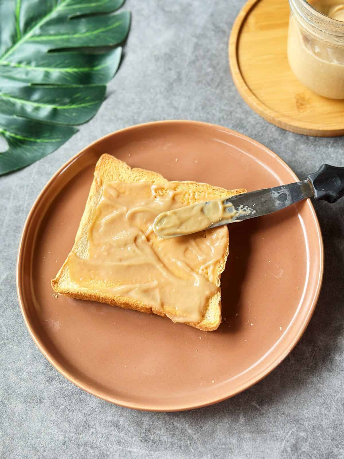 Golden slice of toasted bread generously spread with Hainanese Kaya coconut jam on a brown plate, with the jar of jam in the background.