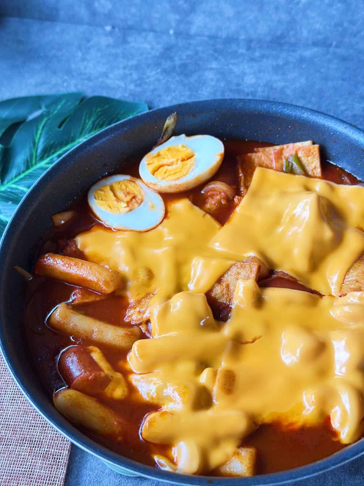 A pan of Korean tteokbokki with chewy rice cakes simmered in a spicy red gochujang sauce, topped with fish cakes, boiled egg, and cheese.