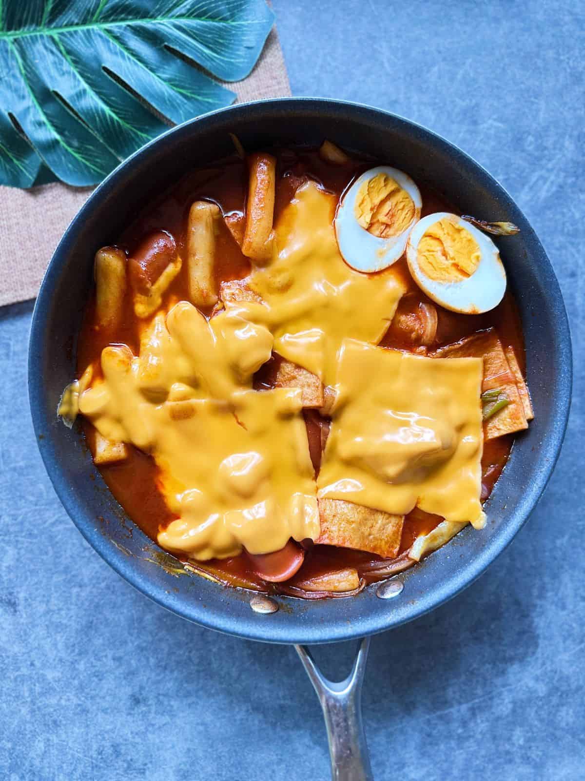 A pan of Korean tteokbokki with chewy rice cakes simmered in a spicy red gochujang sauce, topped with fish cakes, boiled egg, and cheese.