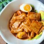 A plate of Korean tteokbokki with chewy rice cakes simmered in a spicy red gochujang sauce, topped with fish cakes, boiled egg, and cheese.
