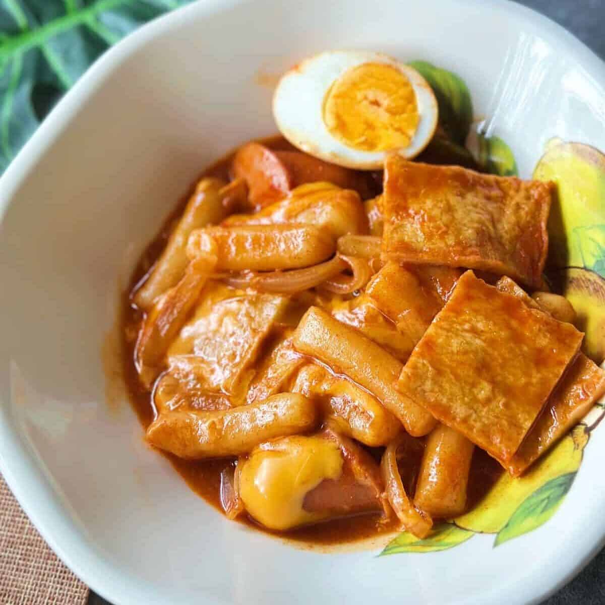 A plate of Korean tteokbokki with chewy rice cakes simmered in a spicy red gochujang sauce, topped with fish cakes, boiled egg, and cheese.