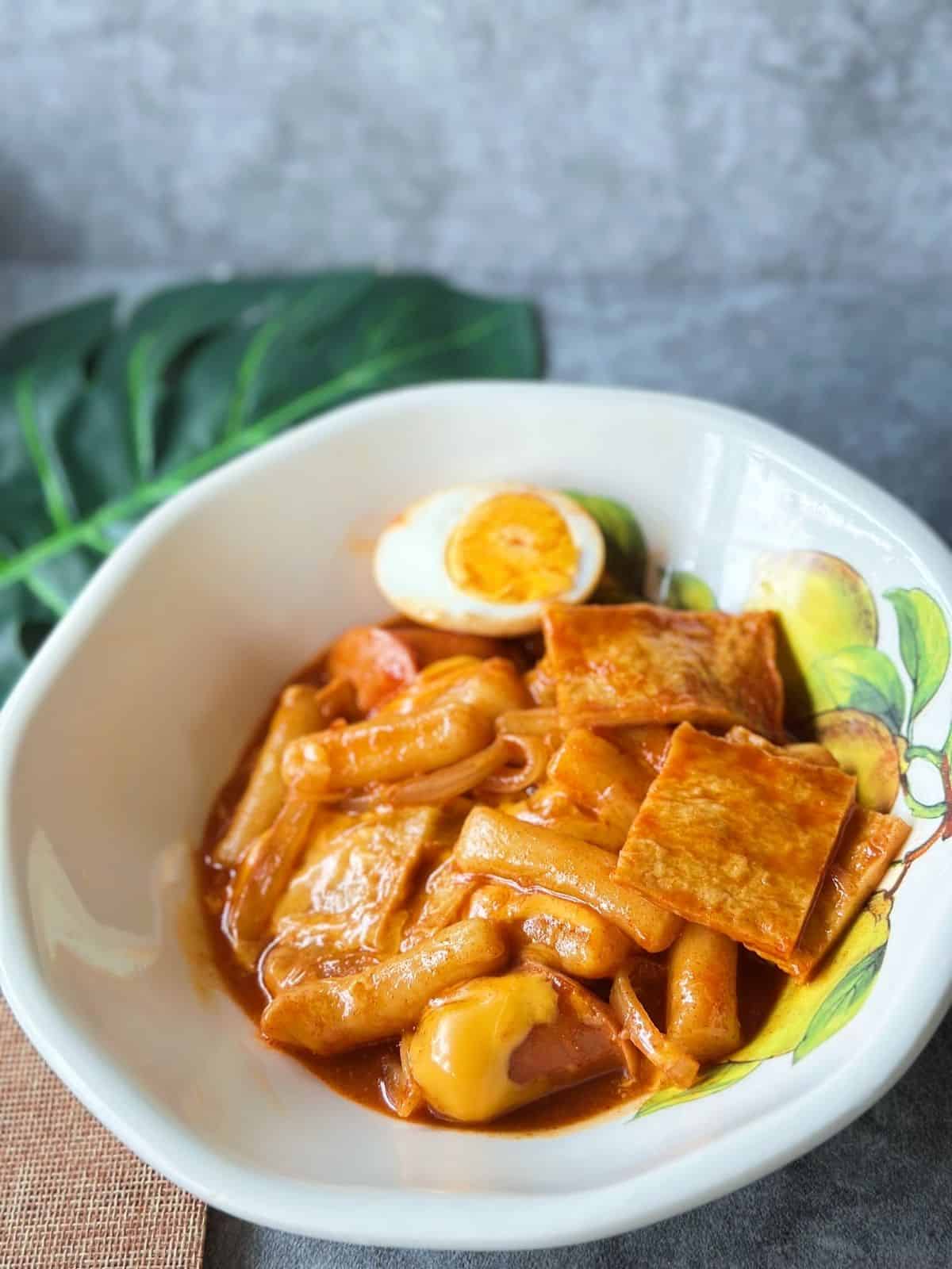 A plate of Korean tteokbokki with chewy rice cakes simmered in a spicy red gochujang sauce, topped with fish cakes, boiled egg, and cheese.