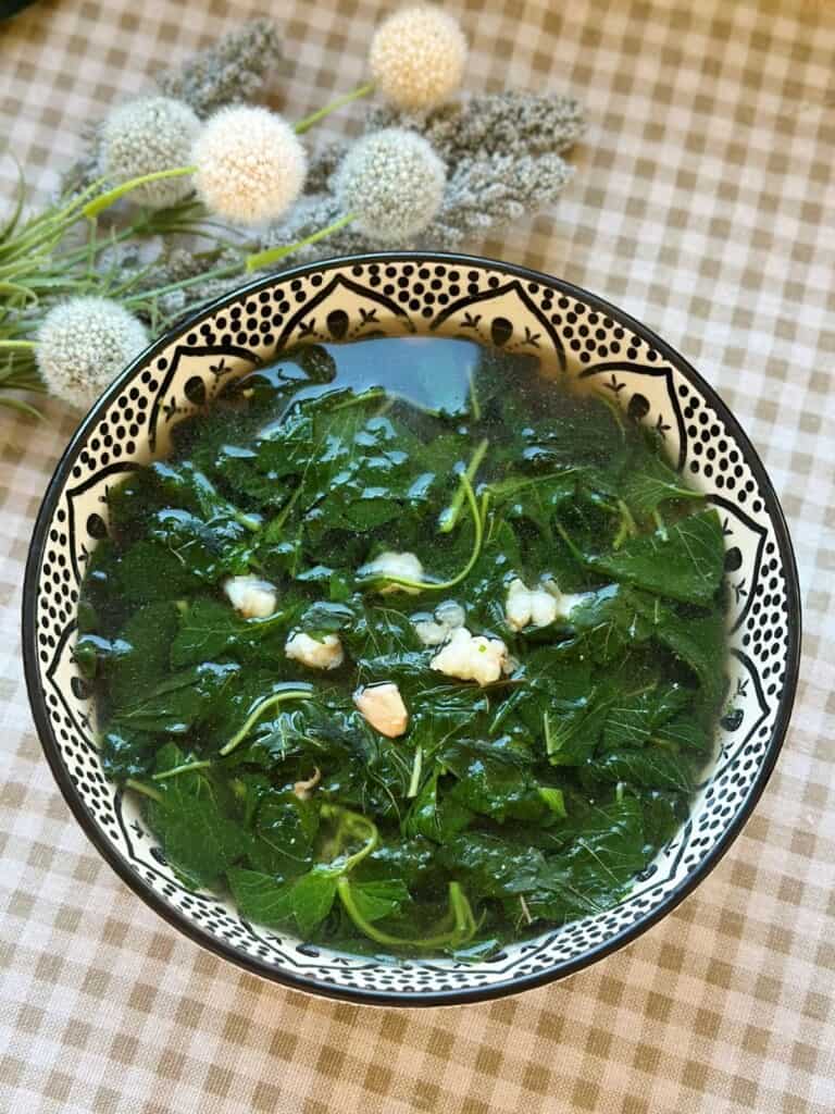 A steaming bowl of Canh Rau Den, Vietnamese amaranth soup, with tender green and red amaranth leaves floating in a clear broth, served simply in a patterned bowl.