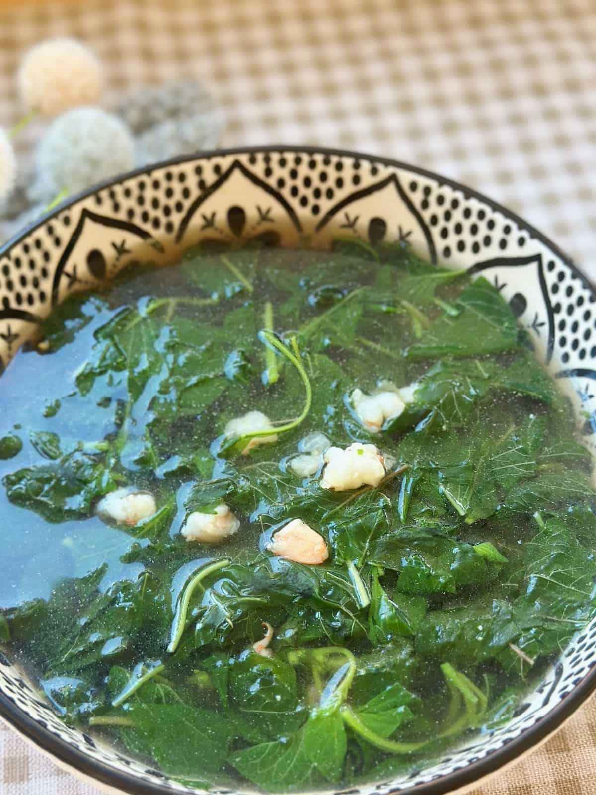 A steaming bowl of Canh Rau Den, Vietnamese amaranth soup, with tender green and red amaranth leaves floating in a clear broth, served simply in a patterned bowl.