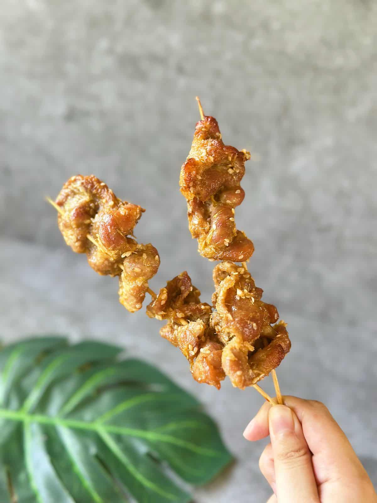 Hand holding two skewers of smoky Vietnamese grilled pork (Thịt Nướng) fresh off the grill
